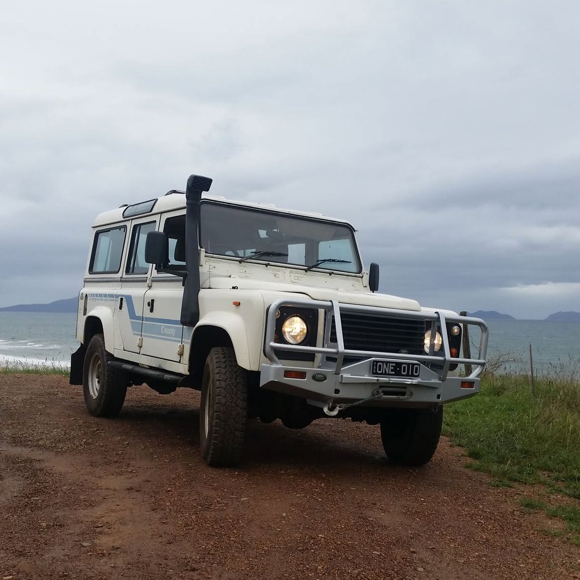 A white Land Rover Defender 110 County with a snorkel and bull bar on a coastal track in Tasmania